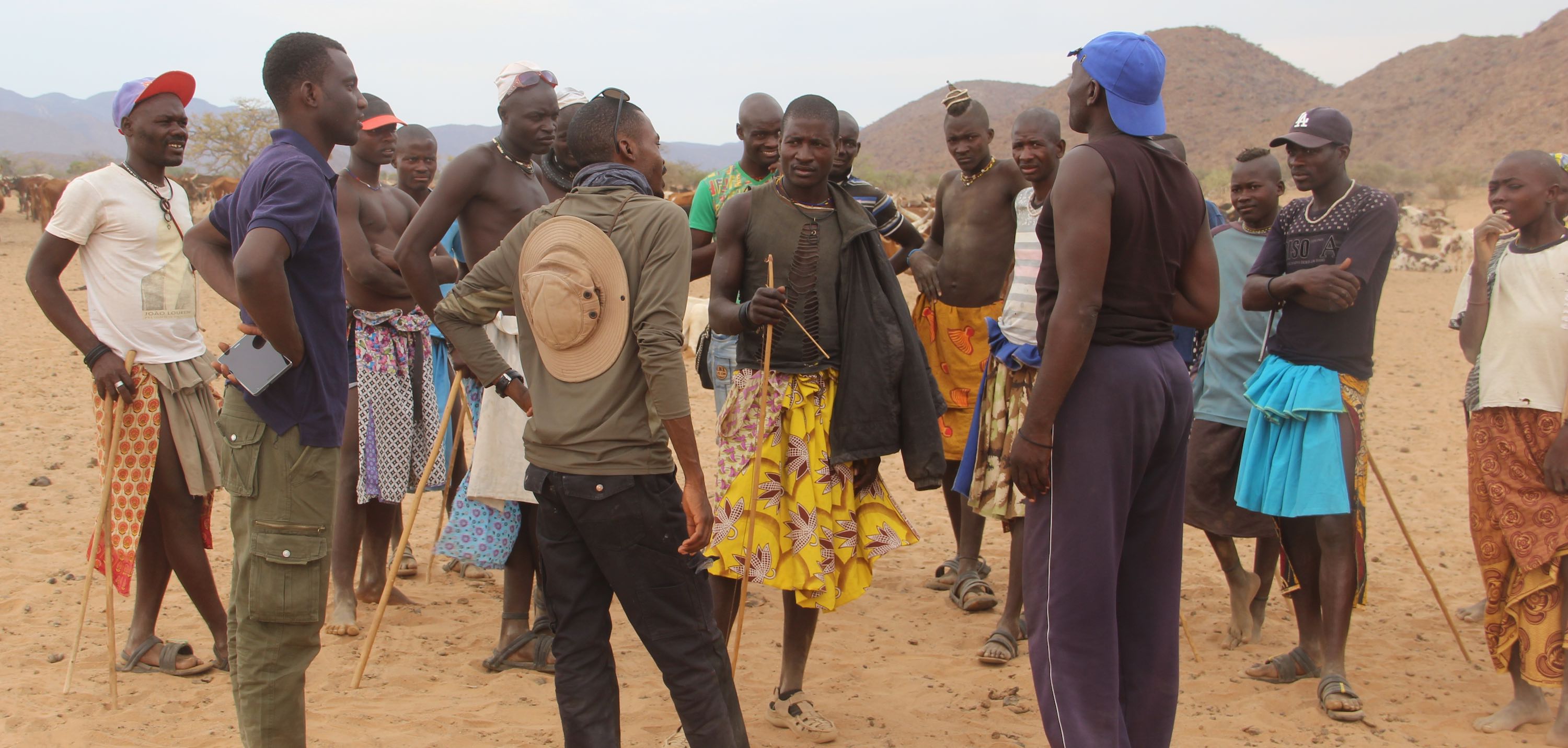 Eighteen people stand in a group on sandy ground, with cows in the background.