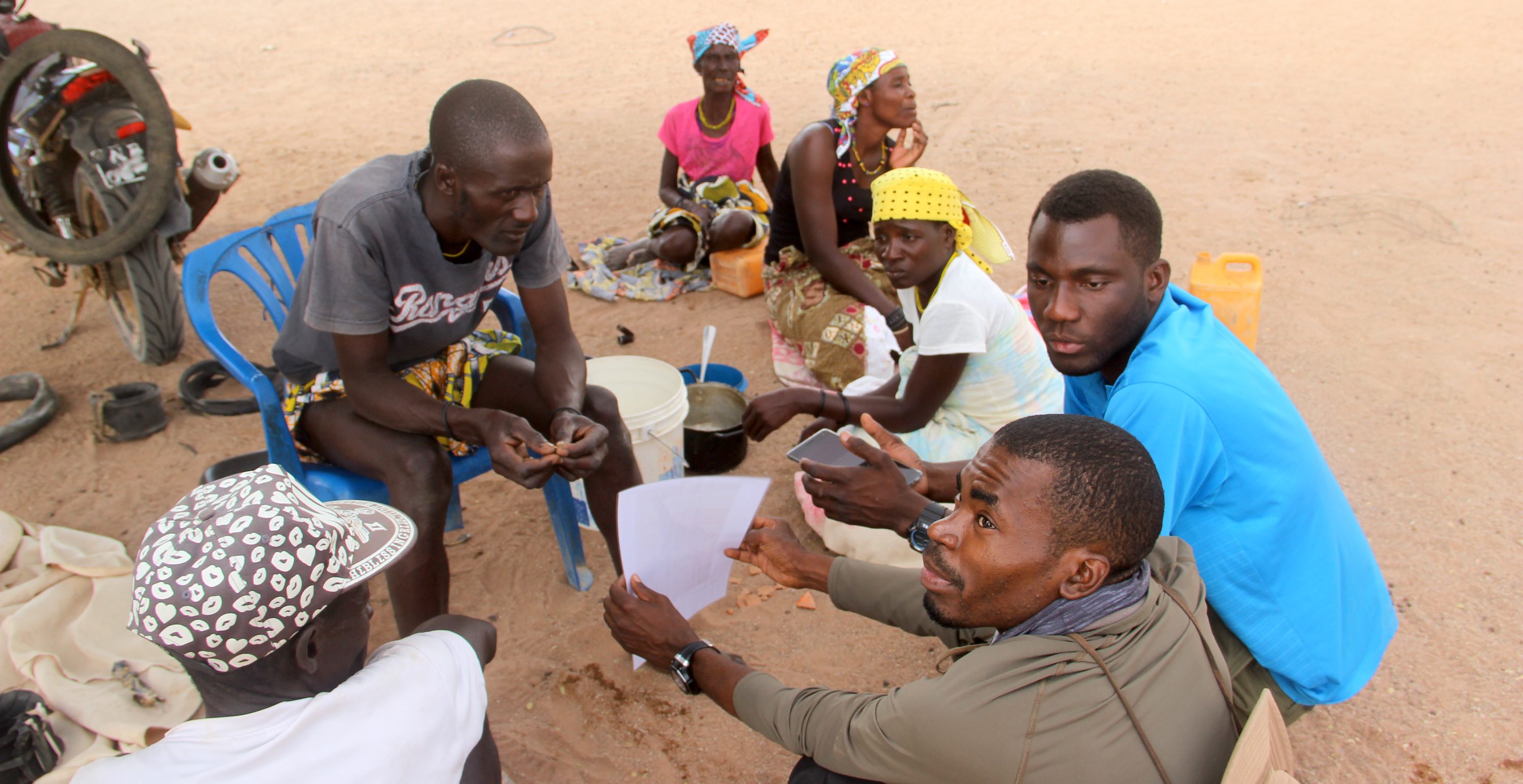 A small group of people sit in a rough circle on the sandy ground.