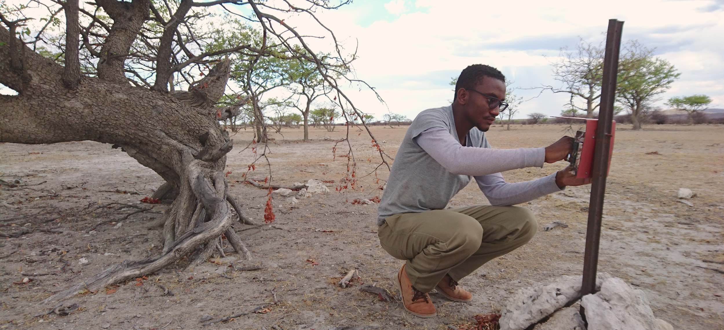 A researcher squats next to a camera trap mounted on a metal pole.