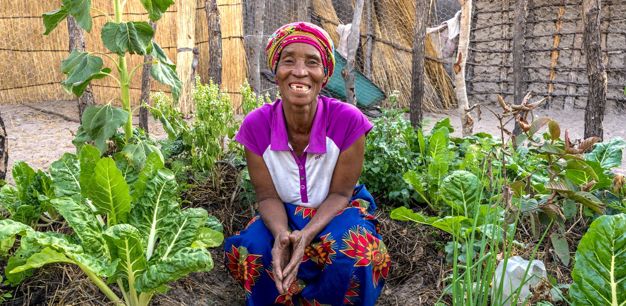 A woman sits smiling on the ground, surrounded by green plants.