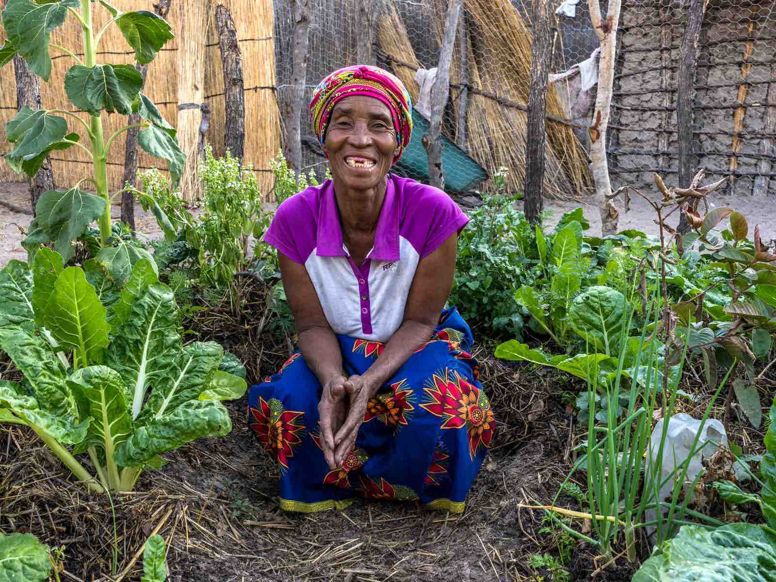 A woman sits smiling on the ground, surrounded by green plants.