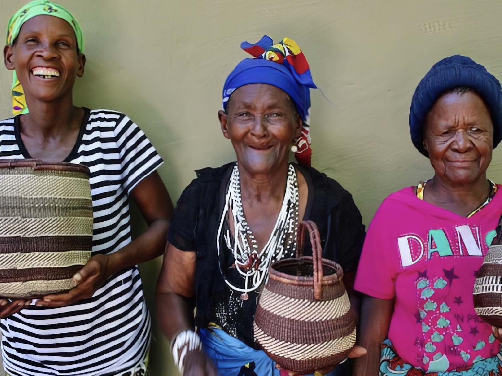 Three colourfully dressed San ladies holding woven baskets and smiling at the camera.